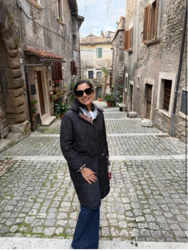 Payal Bhargava, crochet teacher, smiling on a cobbled laneway in a quilted coat and sunglasses