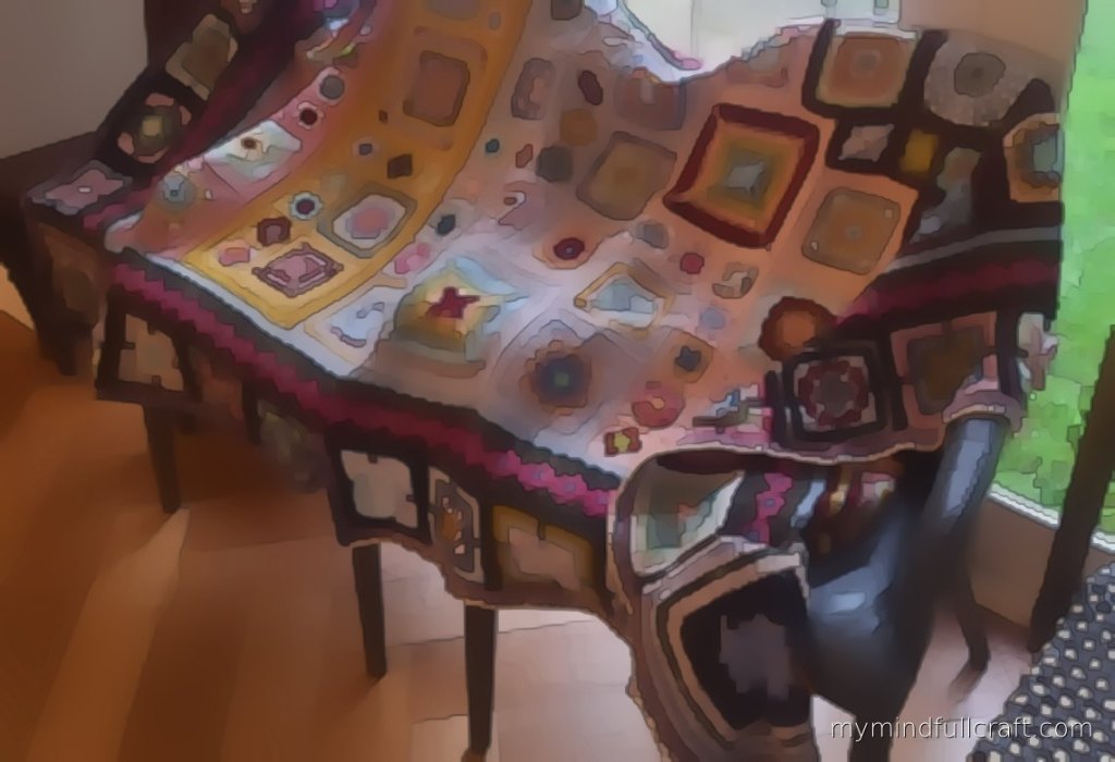 A finished piece from our studio: a crochet sampler blanket in pastel squares and mandalas with a deep plum border, draped over a chair by a window