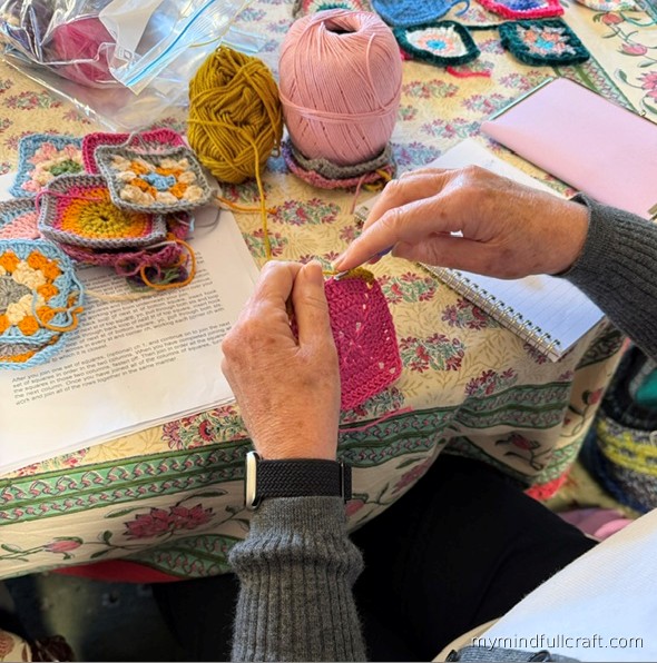 Hands crocheting a pink granny square at a studio table covered in colourful squares and balls of yarn